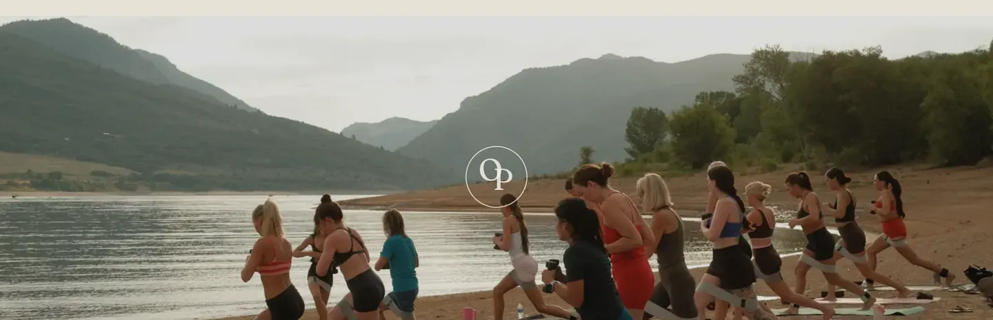 A class of Origin Pilates students lunging on a lake beach below green mountains, with the OP monogram over the water.