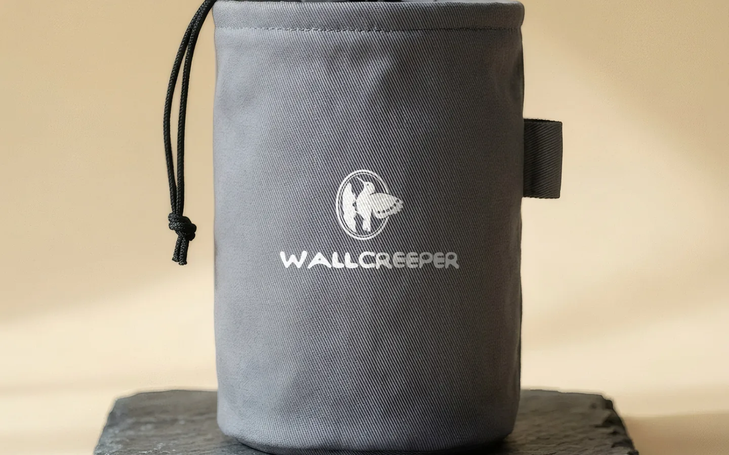 A matte grey twill chalk bag on a slab of slate with the Wallcreeper seal and wordmark printed in chalk white.
